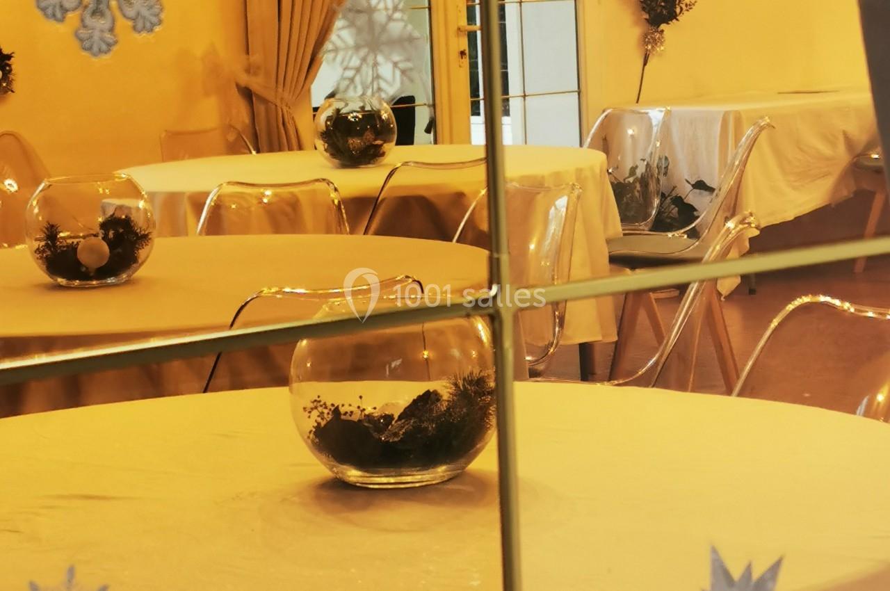 Salle décorée avec des tables rondes, nappes blanches, boules en verre et motifs de flocons sur une vitre.