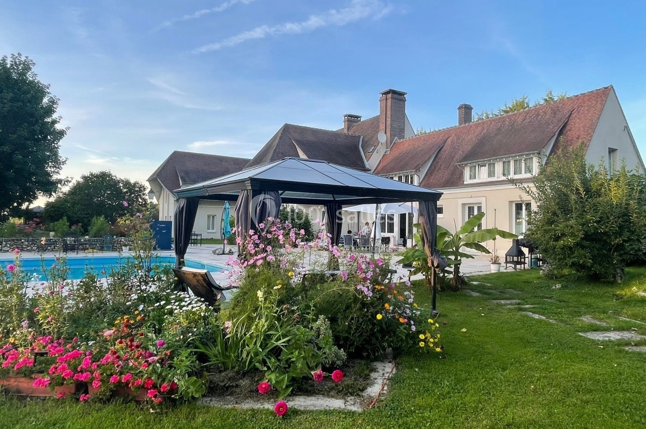Jardin fleuri avec pergola près d'une piscine, devant une grande maison à toit en pente sous un ciel dégagé.
