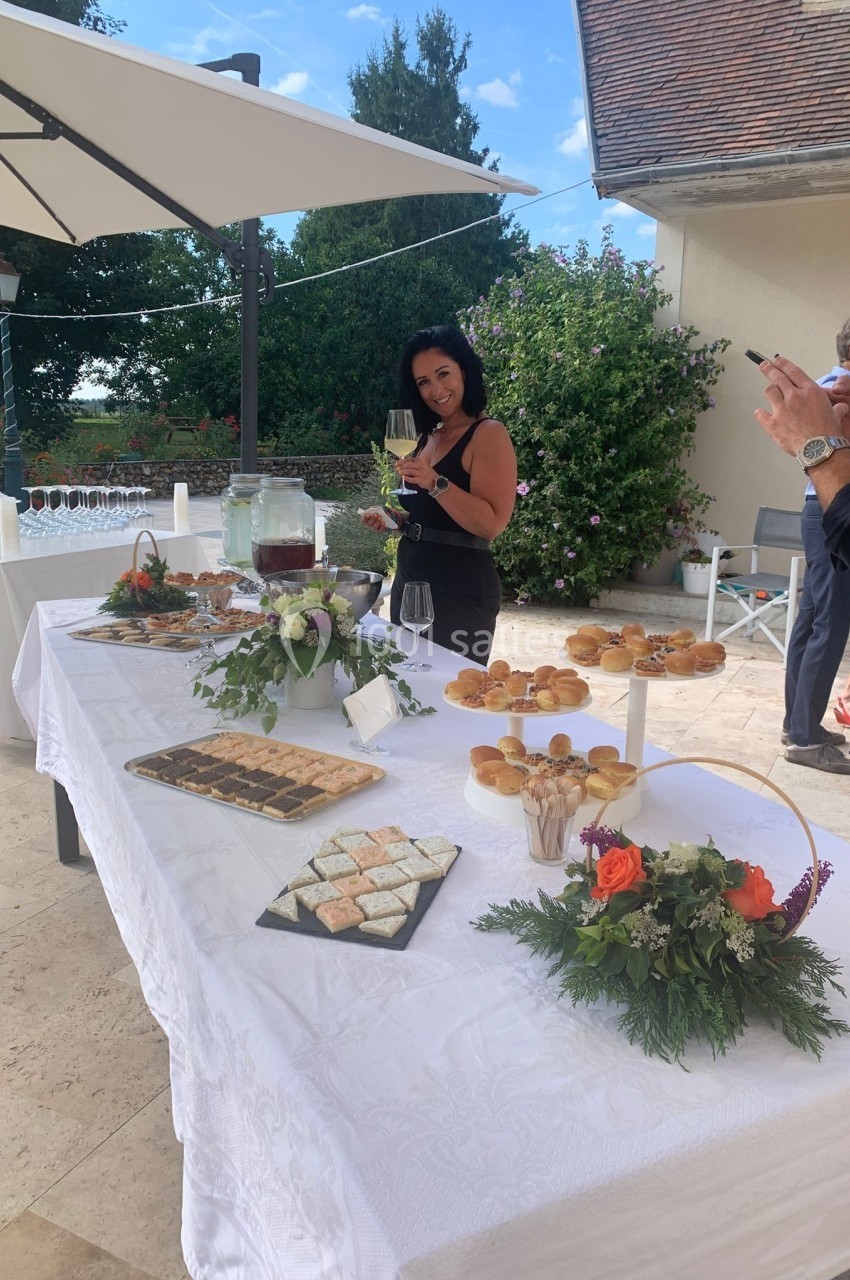 Table dressée en extérieur avec des amuse-bouches, une femme tenant un verre et un homme prenant une photo.