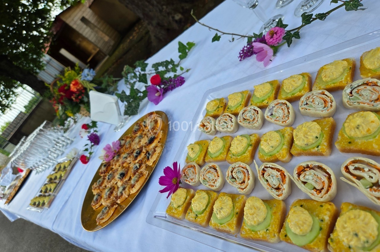 Plateau de bouchées apéritives variées disposé sur une table décorée de fleurs, avec verres et serviettes en arrière-plan.