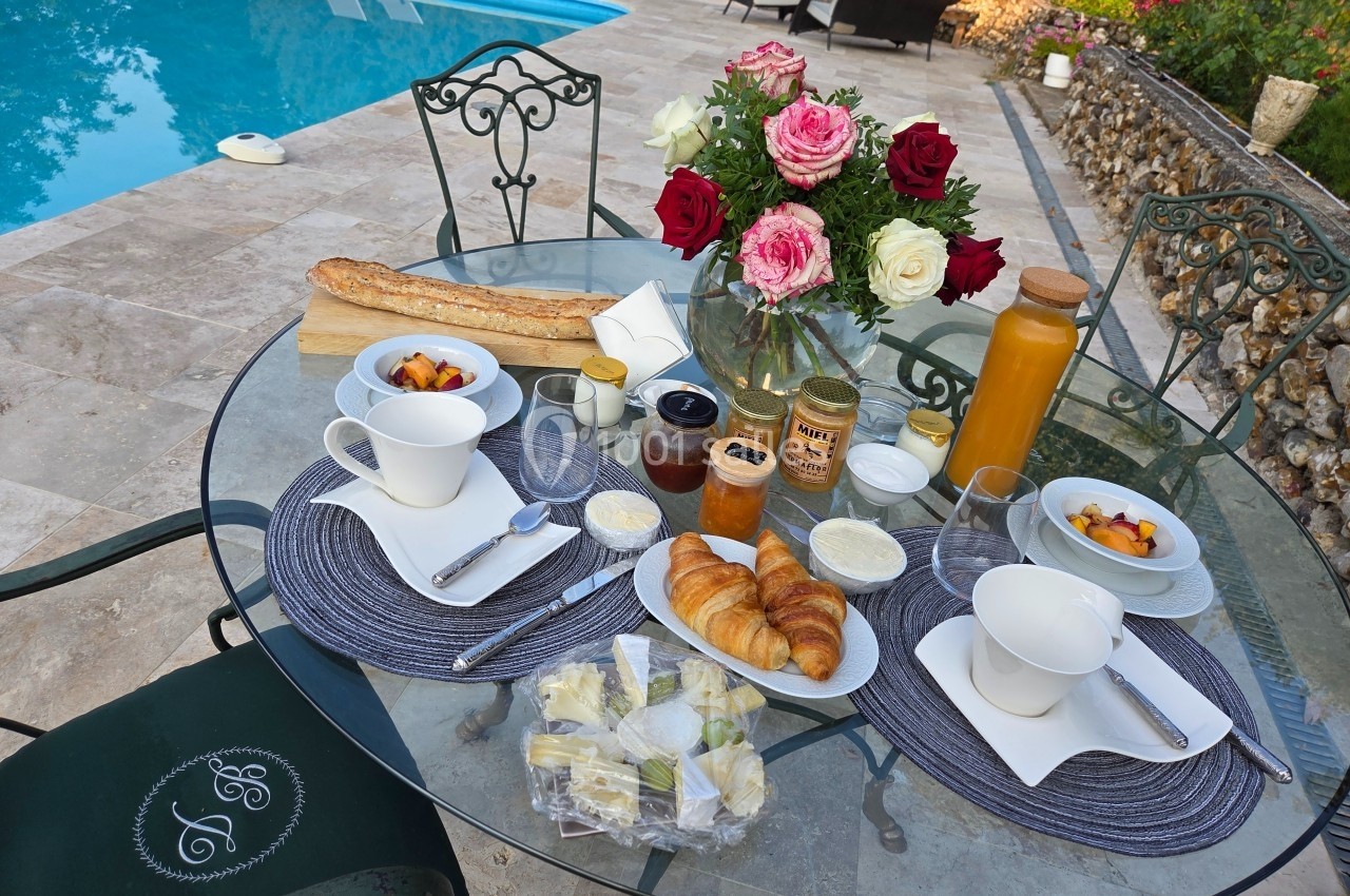 Petit-déjeuner servi en extérieur sur une table ronde près d'une piscine, avec viennoiseries, fruits, jus et fleurs.