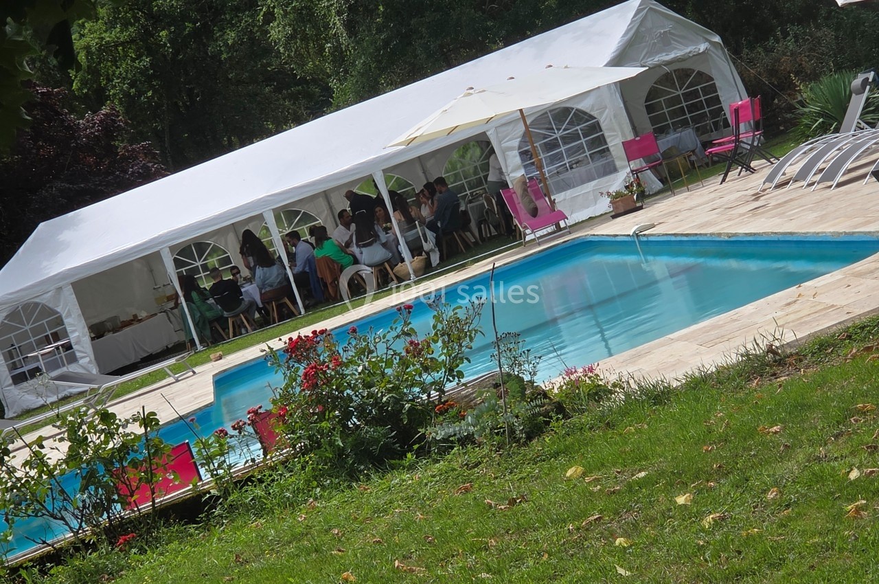 Tente blanche installée près d'une piscine entourée de chaises longues, avec des personnes assises à l'intérieur.