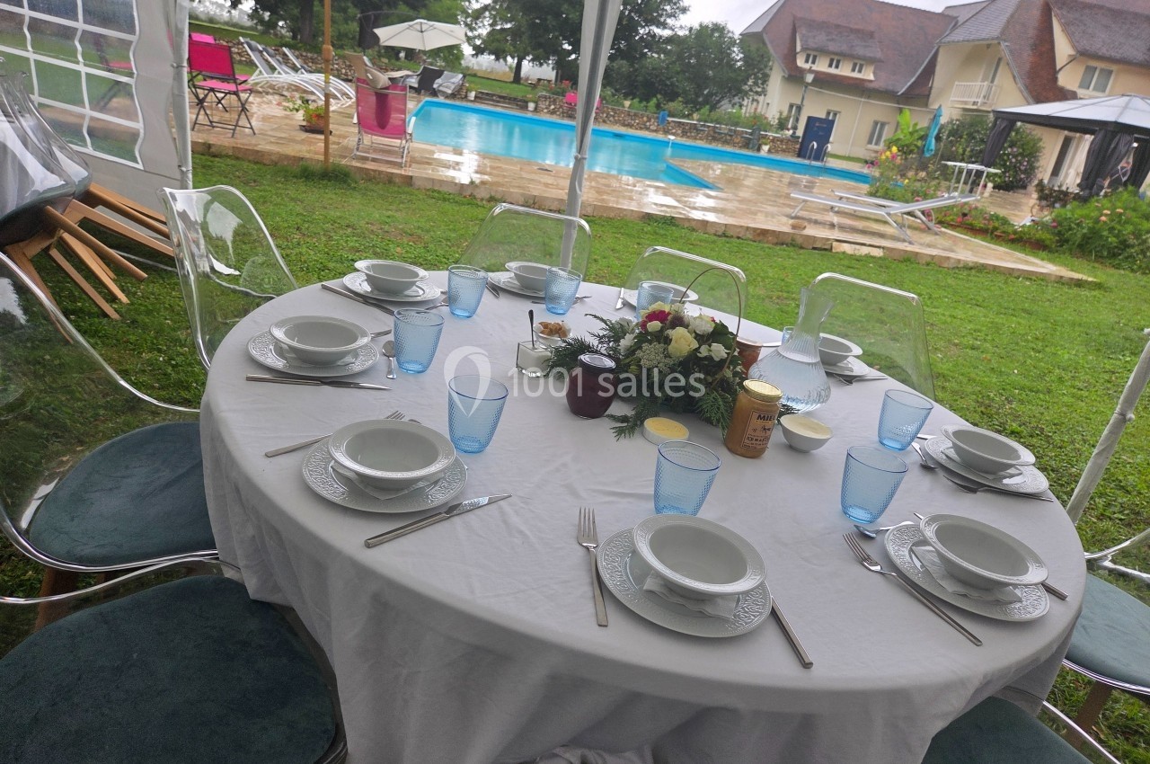 Table dressée avec vaisselle blanche et verres bleus sous une tente, près d'une piscine et d'un jardin verdoyant.