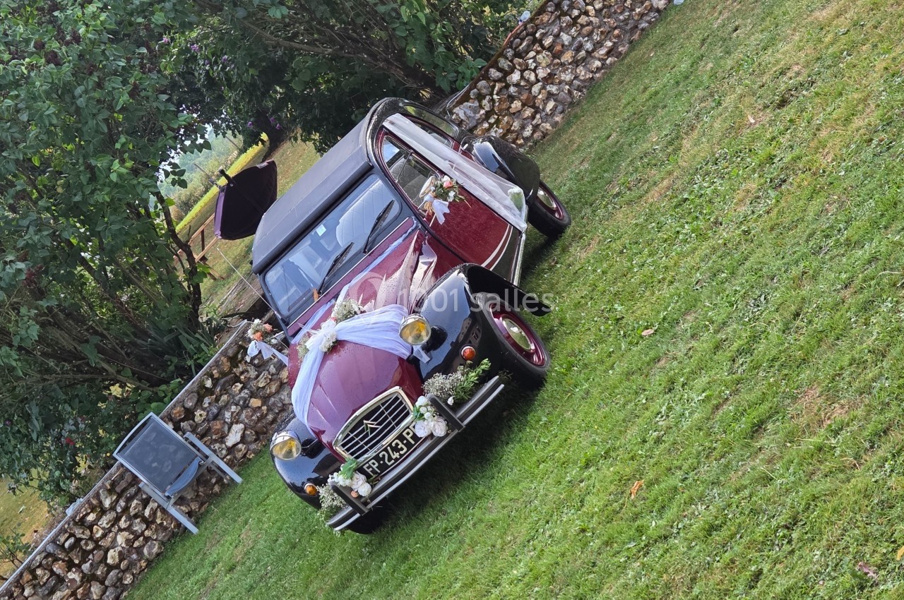 Voiture ancienne décorée pour un mariage, stationnée sur une pelouse près d'un muret en pierre.