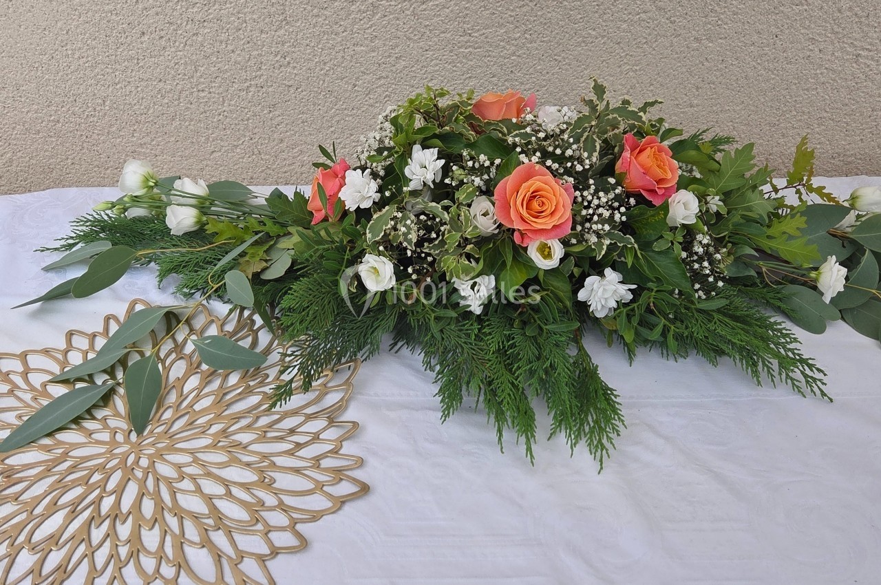 Composition florale avec roses orange, fleurs blanches et feuillage vert, disposée sur une nappe blanche.