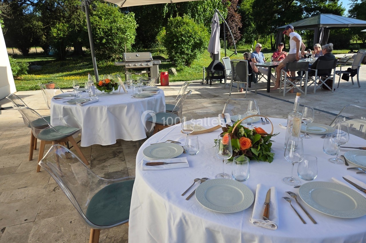 Tables dressées avec nappes blanches et décoration florale sur une terrasse ensoleillée, avec des convives en arrière-plan.