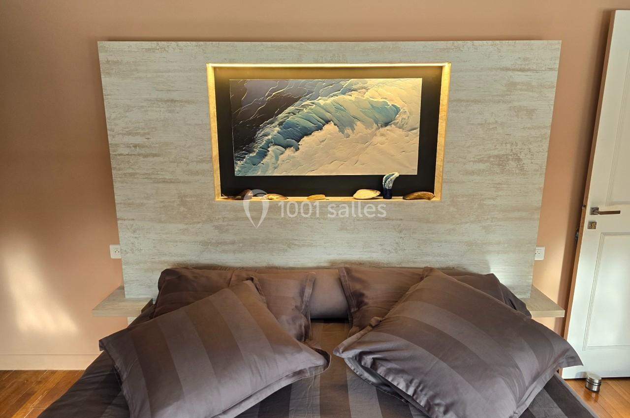 Lit avec des coussins gris devant une tête de lit en bois clair, ornée d'un tableau de vague éclairé.