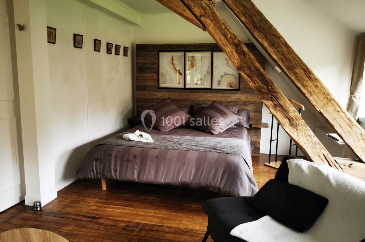 Chambre mansardée avec poutres apparentes, lit double, coussins violets, et fauteuil noir sur un parquet en bois.