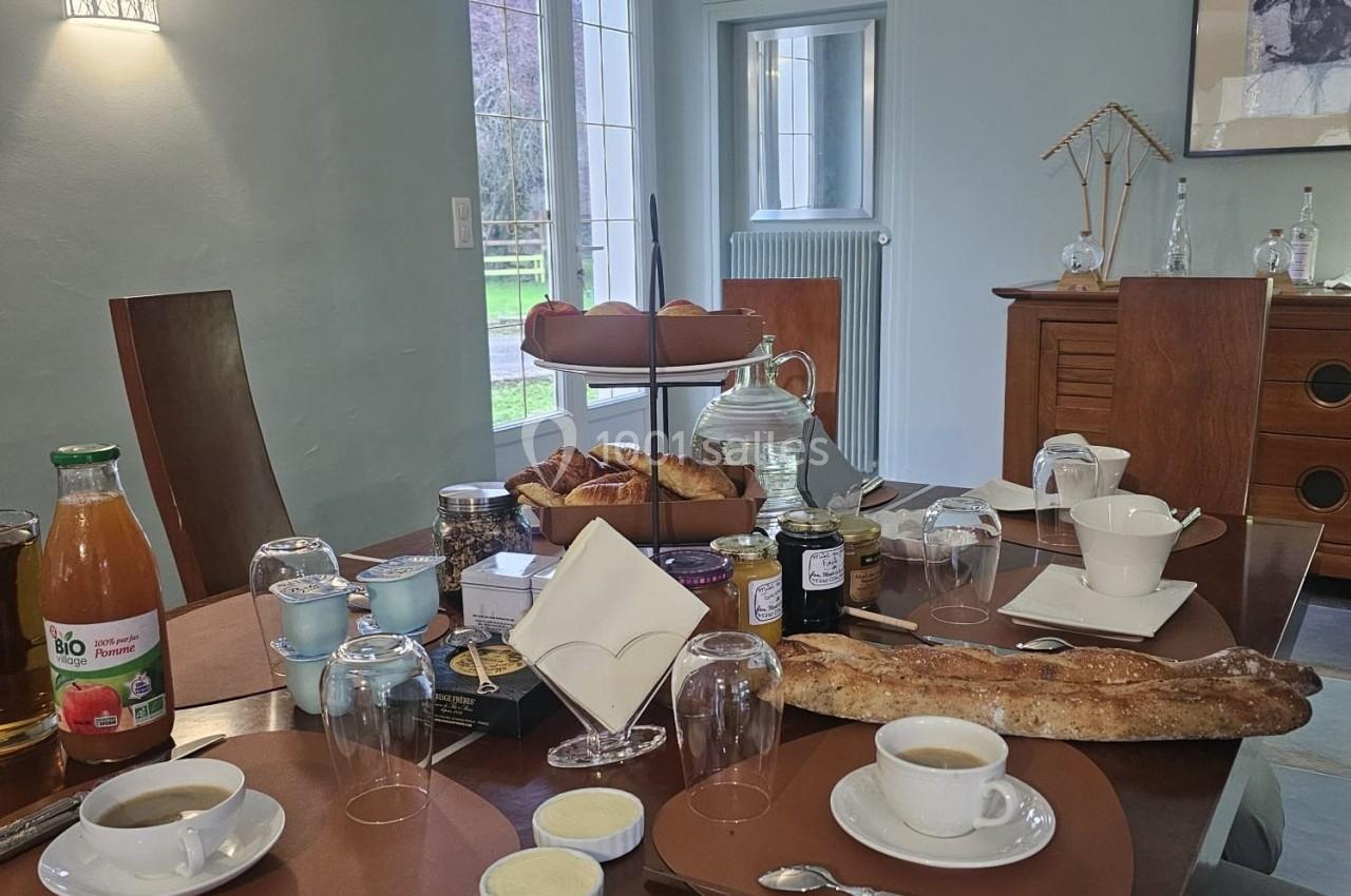 Table dressée pour un petit-déjeuner avec pain, viennoiseries, boissons, confitures et fromages dans une salle lumineuse.