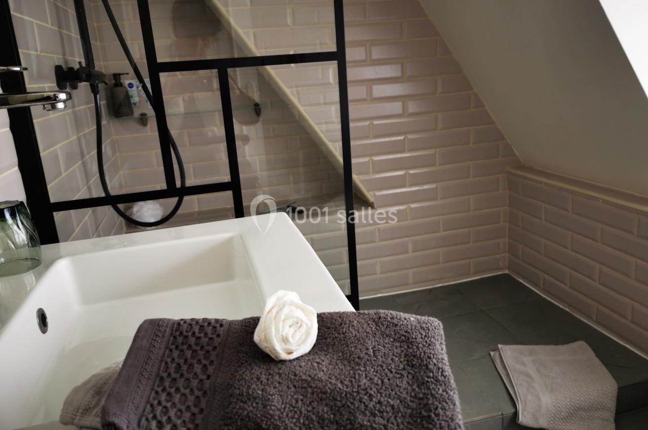 Salle de bain mansardée avec douche à paroi vitrée, lavabo blanc et serviettes pliées sur le bord.