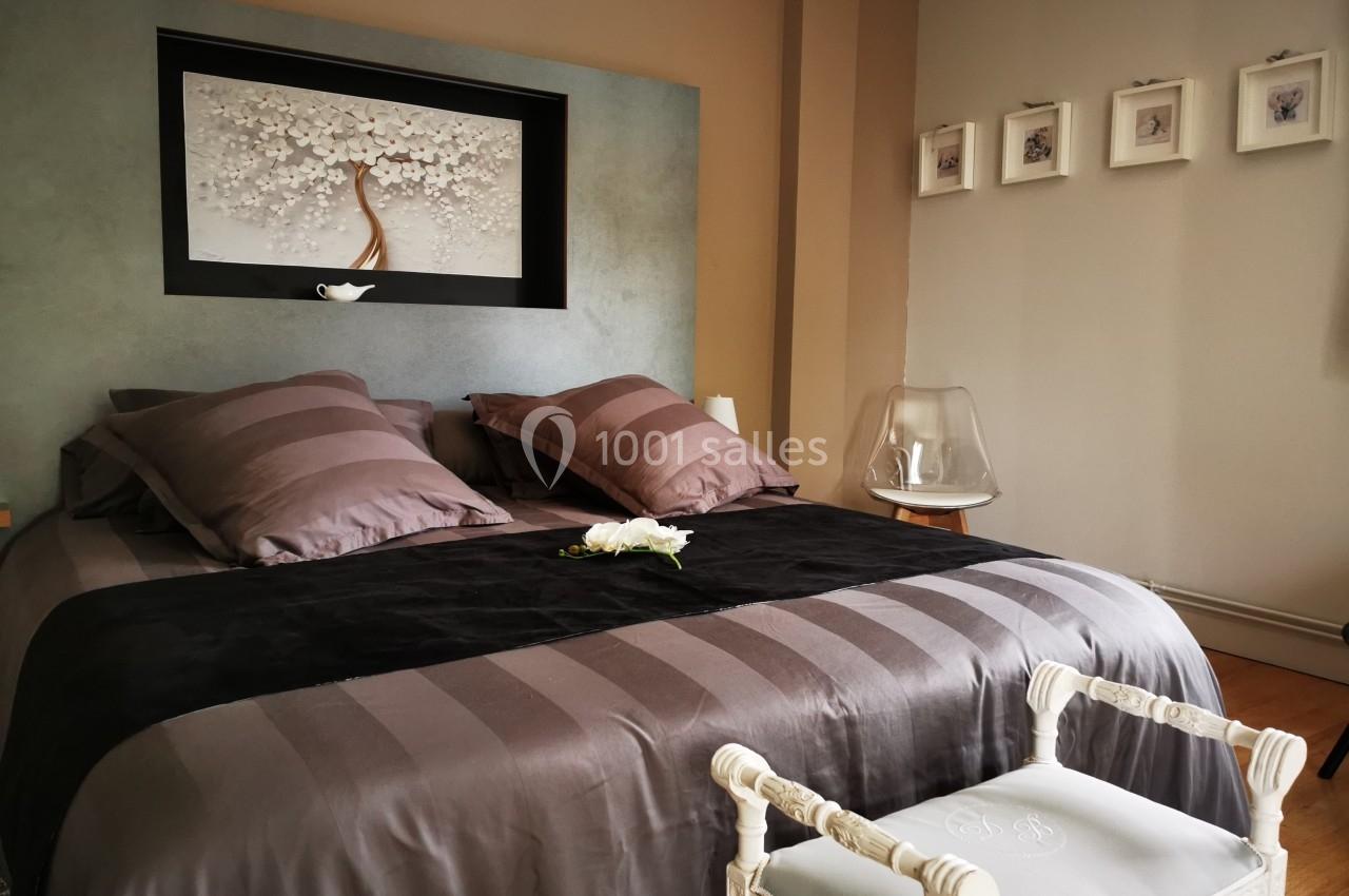 Lit double avec couvre-lit gris, coussins assortis, tableau floral au mur et chaise blanche au pied du lit.