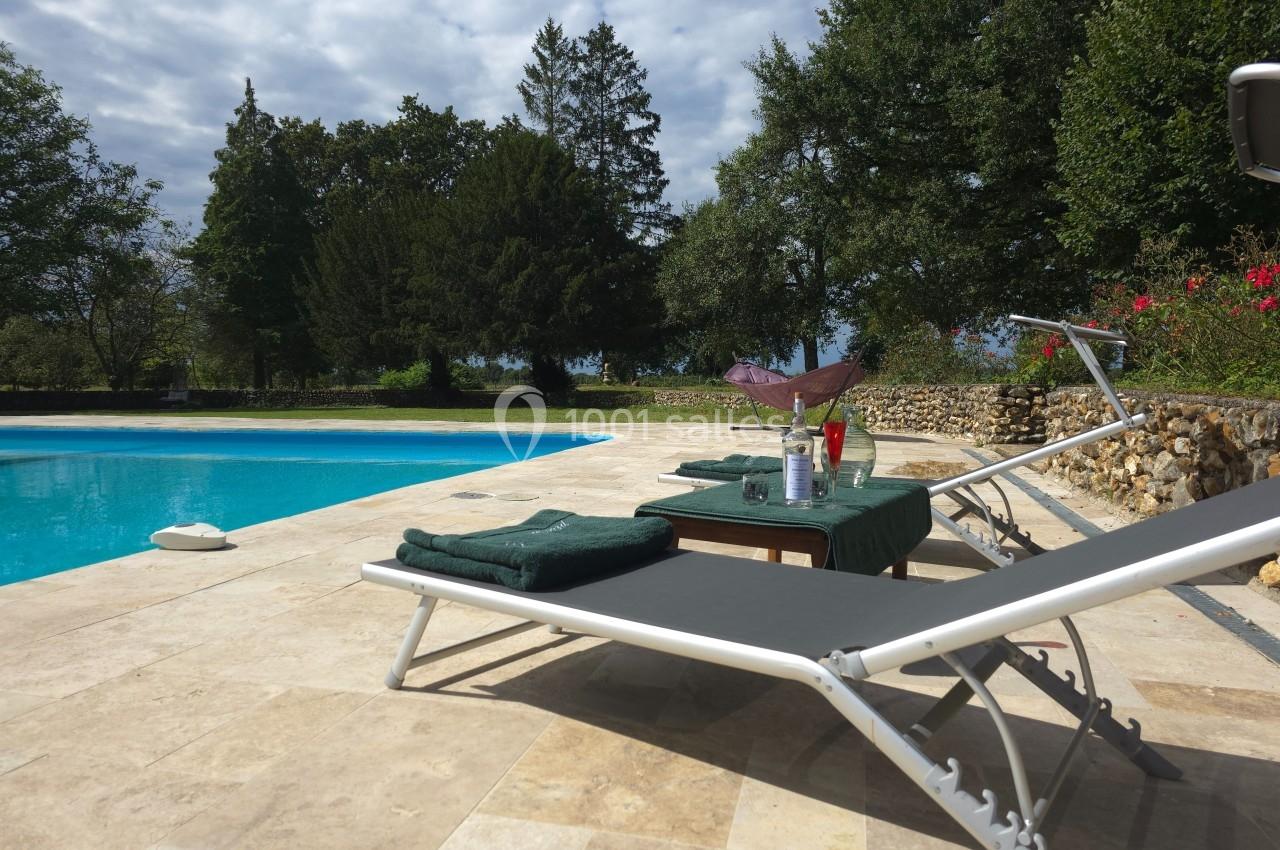 Deux transats près d'une piscine, avec une table dressée contenant des verres et une bouteille, entourée de végétation.