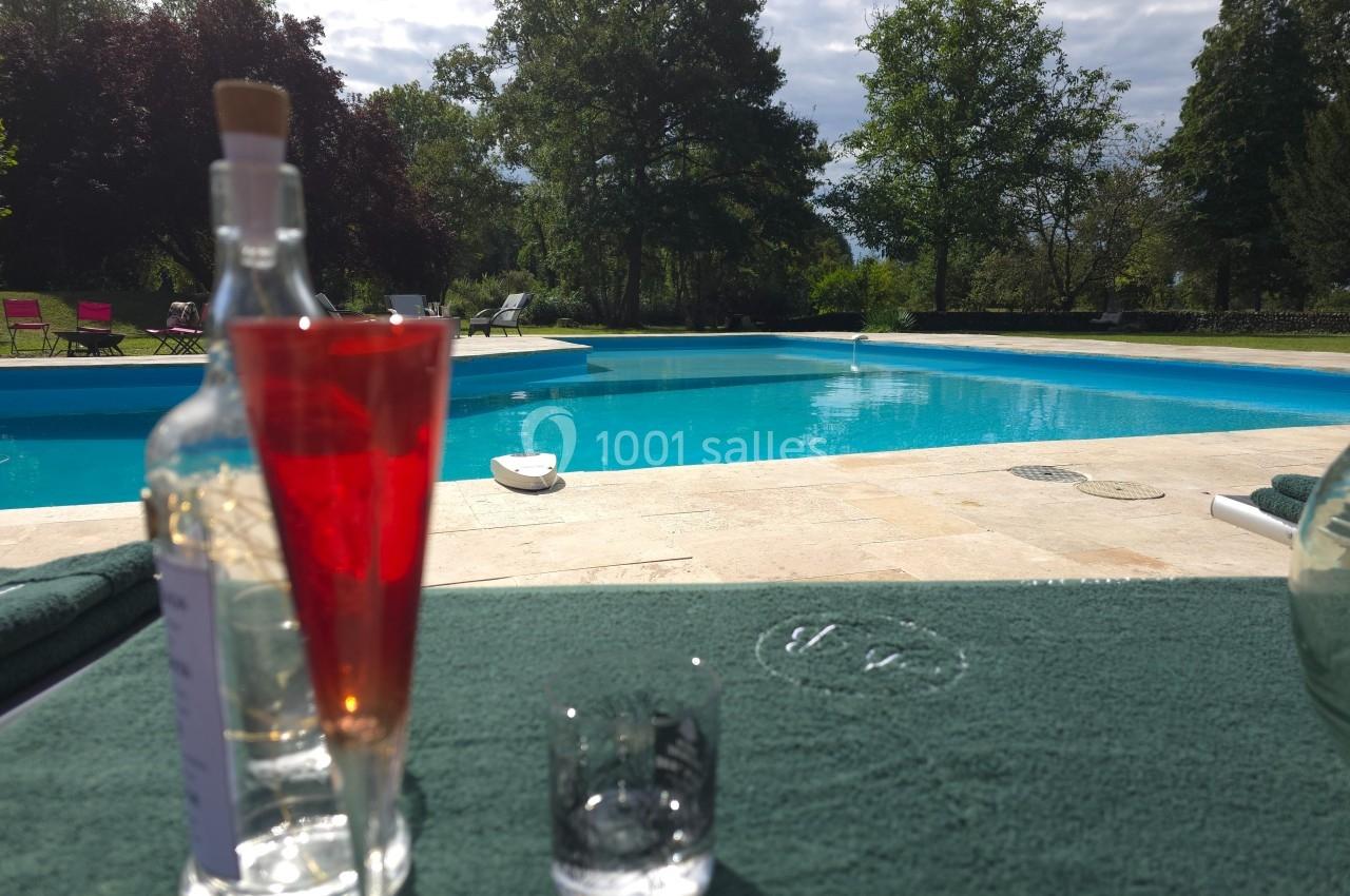 Vue d'une piscine extérieure entourée d'arbres, avec des verres et une bouteille posés sur une table au premier plan.
