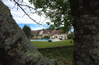 Piscine extérieure bordée de dalles, devant une grande maison traditionnelle avec toit en tuiles rouges.