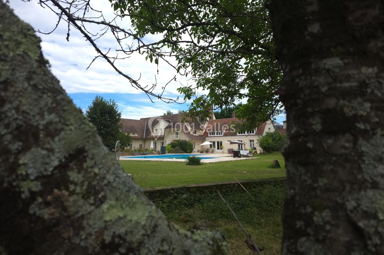 Vue d'une maison avec piscine et jardin, encadrée par les branches d'un arbre au premier plan.