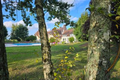 Piscine extérieure bordée de dalles, devant une grande maison traditionnelle avec toit en tuiles rouges.