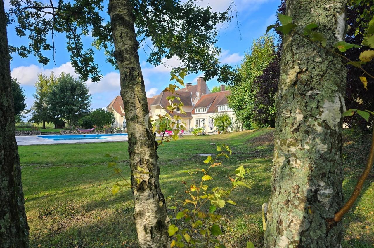 Vue d'une maison entourée d'arbres, avec une pelouse et une piscine sous un ciel bleu.