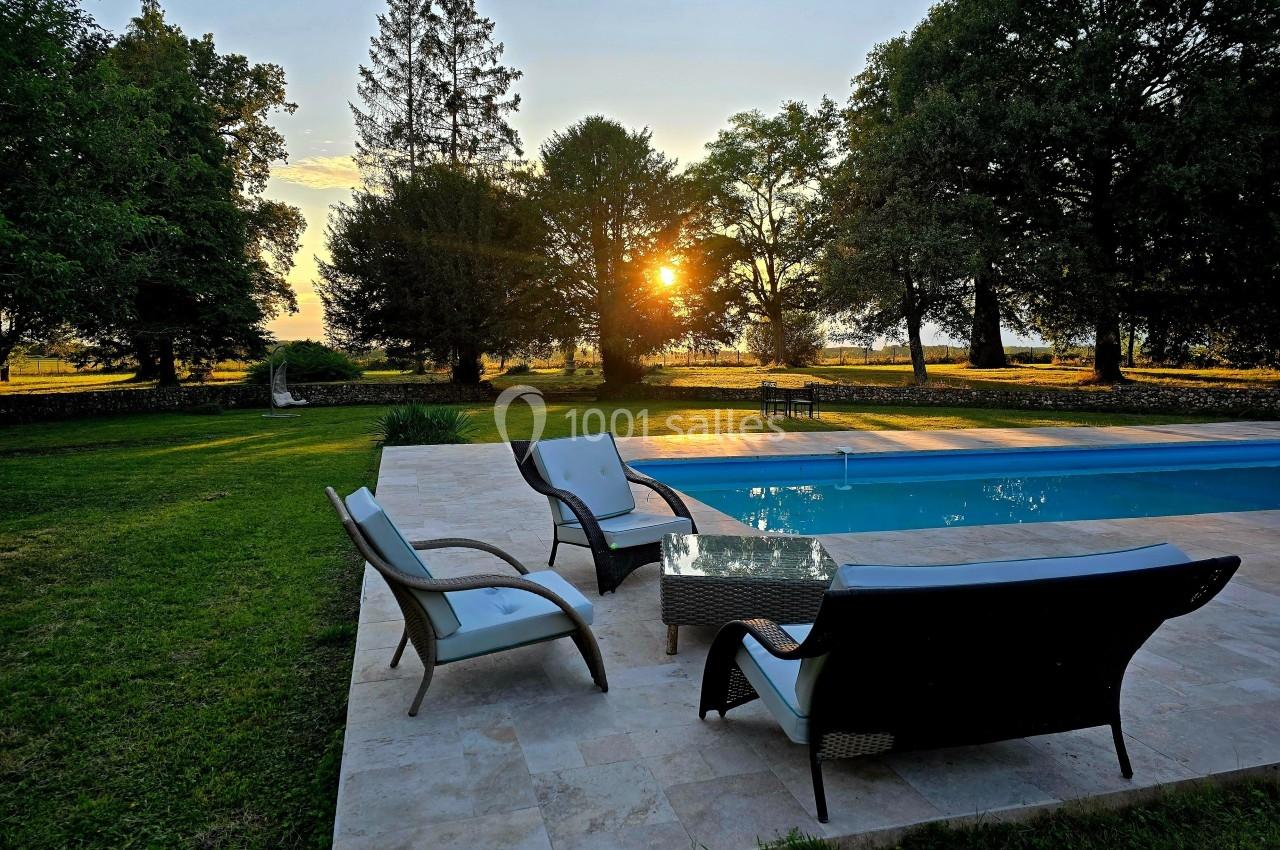Terrasse avec fauteuils et table basse près d'une piscine, entourée de pelouse et d'arbres au coucher du soleil.