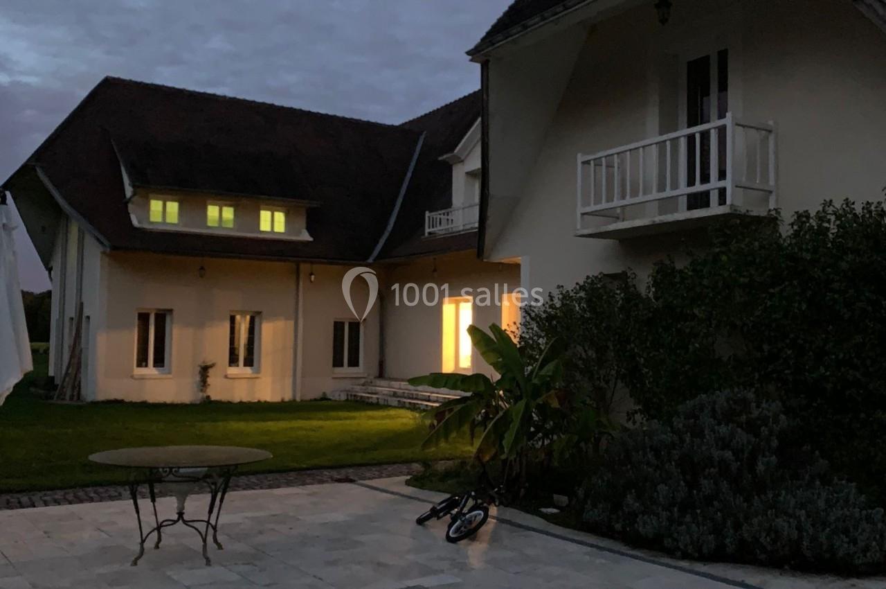 Maison avec toit en pente, éclairée de l'intérieur, vue depuis une terrasse en pierre au crépuscule.