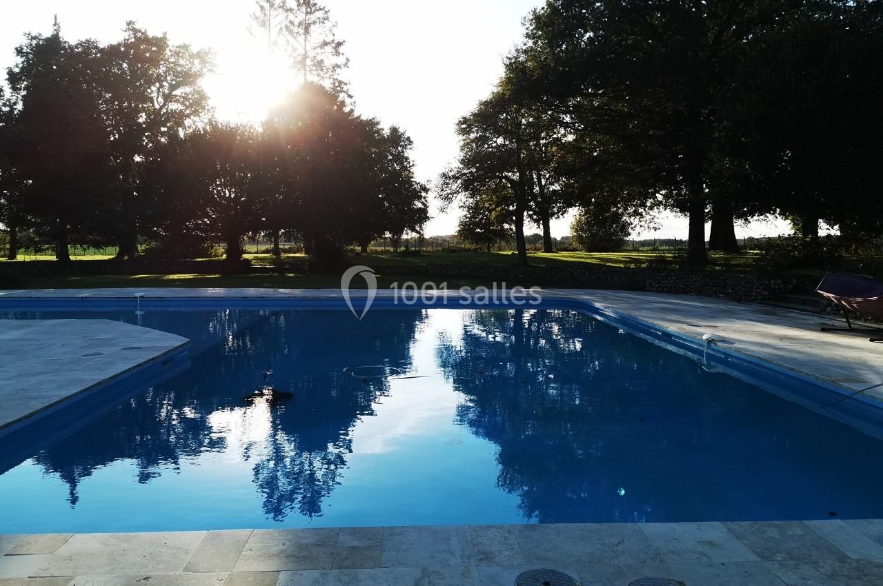 Piscine extérieure entourée d'arbres, reflétant le ciel et le soleil couchant à l'arrière-plan.
