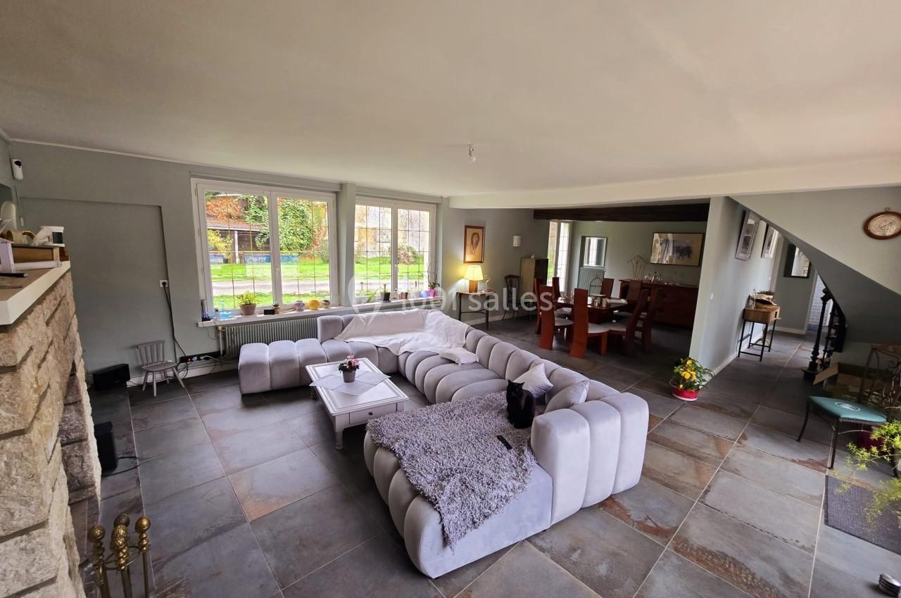 Salon spacieux avec canapé en angle, table basse, grandes fenêtres donnant sur un jardin et coin salle à manger au fond.