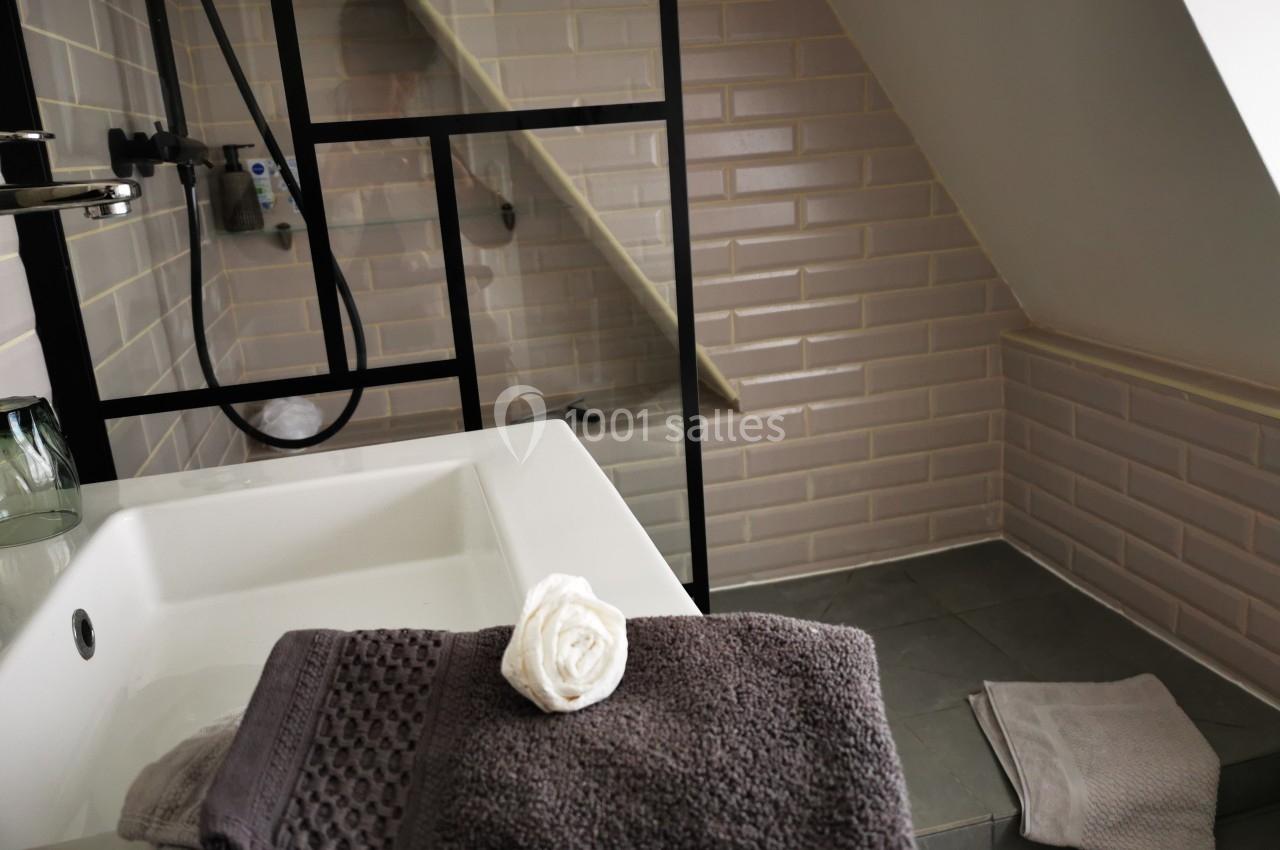 Salle de bain mansardée avec lavabo, serviettes pliées, douche à paroi vitrée et carrelage beige.