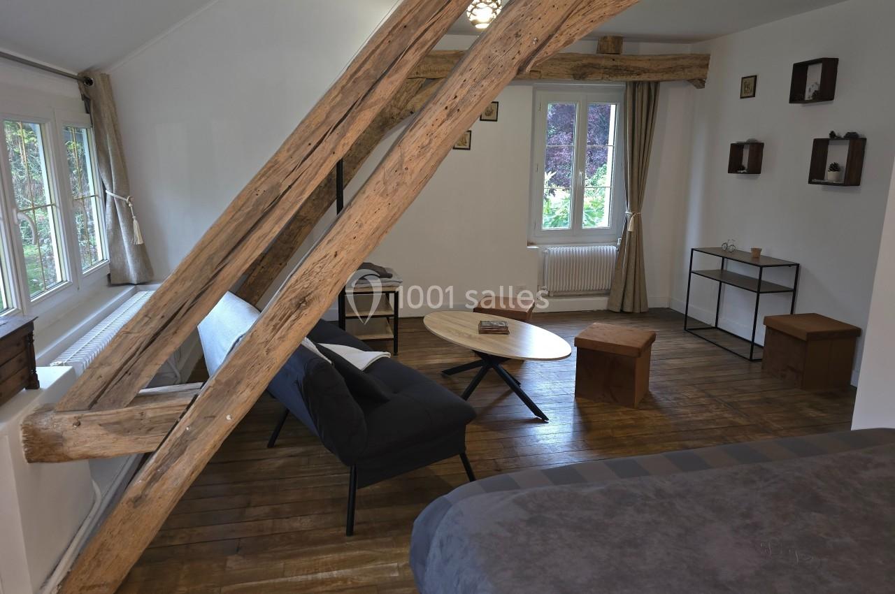 Salon mansardé avec poutres apparentes, canapé, table basse, étagères murales et vue sur un jardin par la fenêtre.