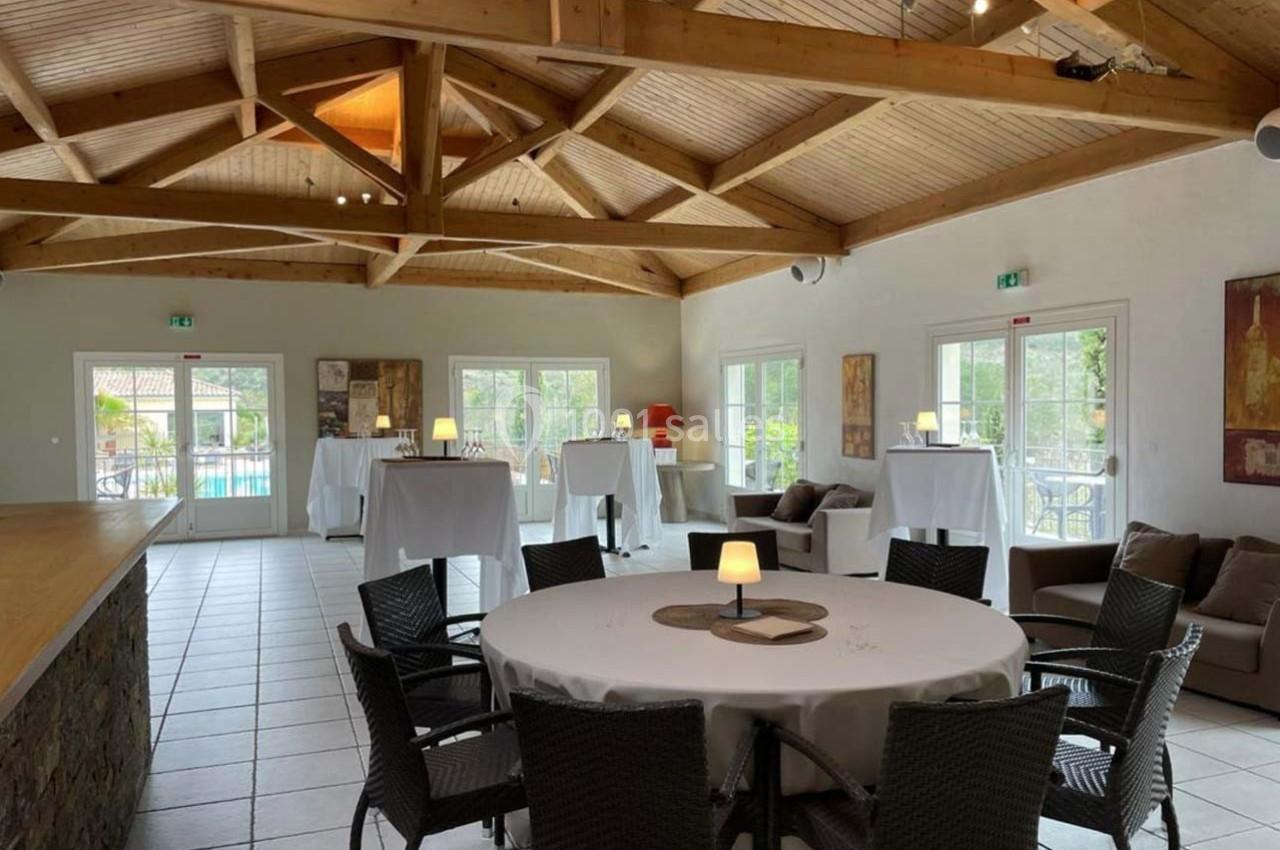 Salle lumineuse avec charpente en bois, tables rondes et hautes nappées de blanc, grandes baies vitrées donnant sur l…