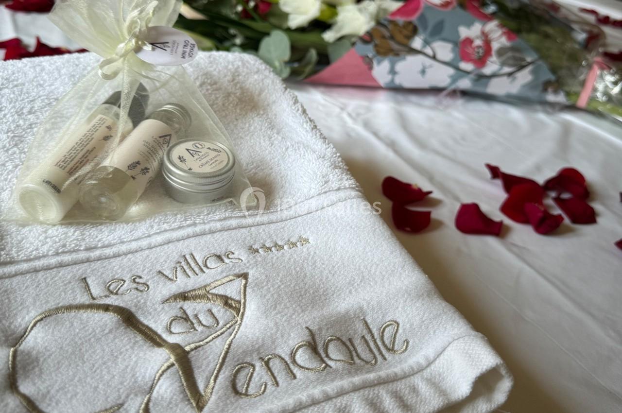 Serviette brodée avec des produits de soin, pétales de rose et bouquet de fleurs en arrière-plan.