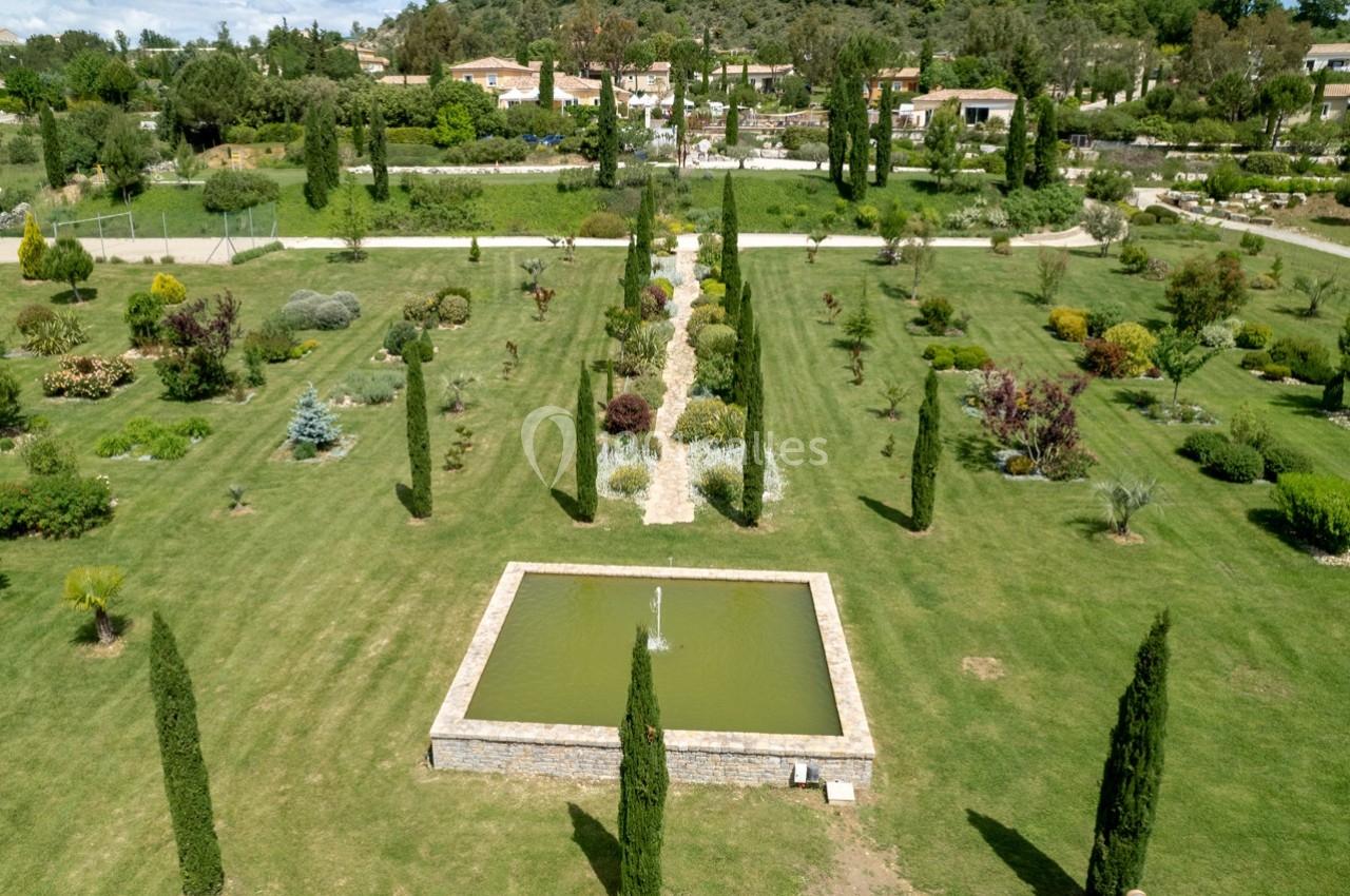 Vue aérienne d'un jardin paysager avec une fontaine centrale, des allées bordées de cyprès et des arbustes variés.