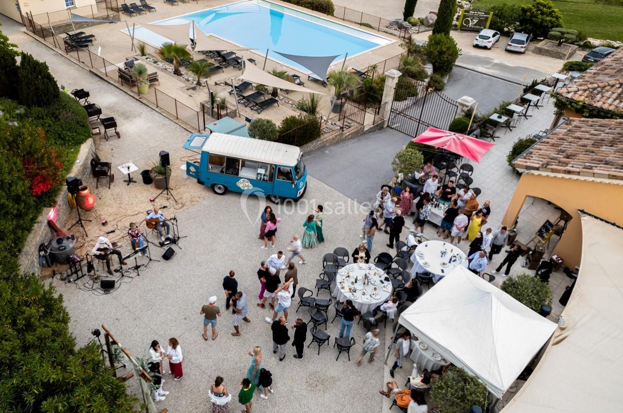 Vue aérienne d'une réception en plein air avec des invités, un food truck, des tables dressées et une piscine en arrière…