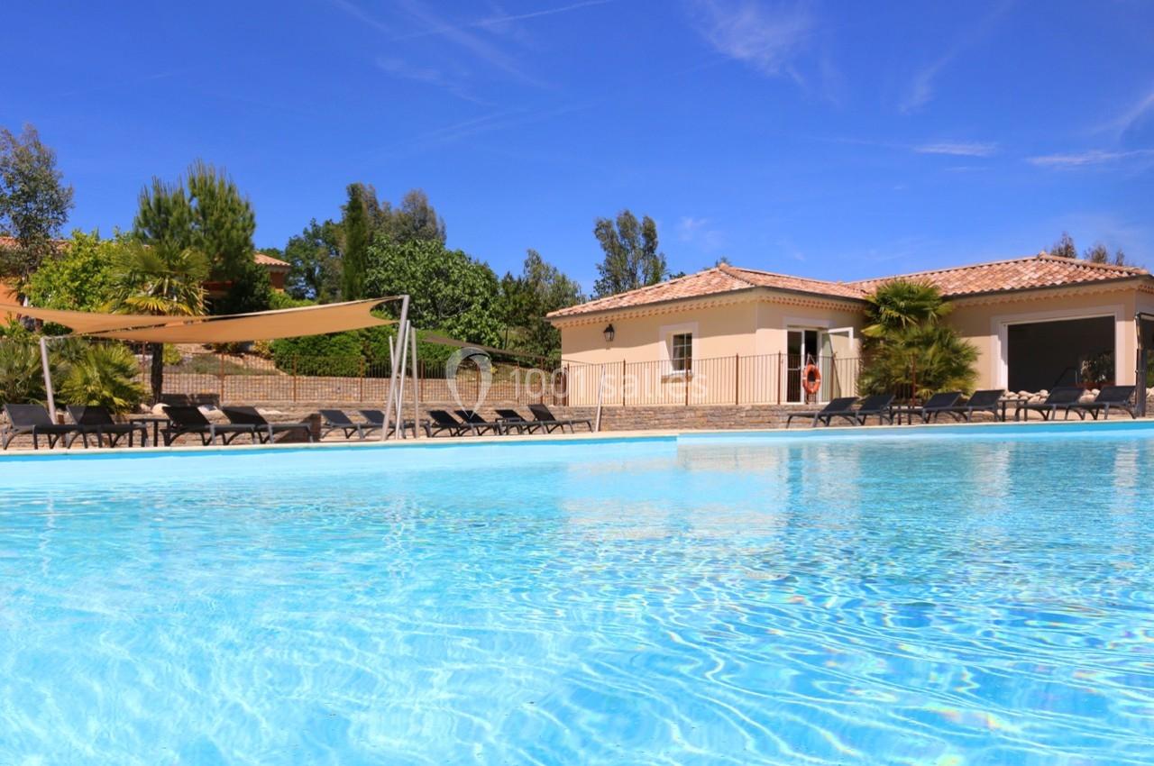 Piscine extérieure avec eau claire, entourée de transats, d'arbres et d'une maison sous un ciel bleu.