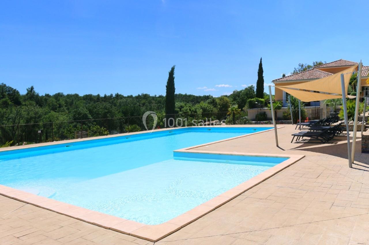 Piscine extérieure entourée de chaises longues et d'une pergola, avec vue sur un paysage verdoyant sous un ciel bleu.