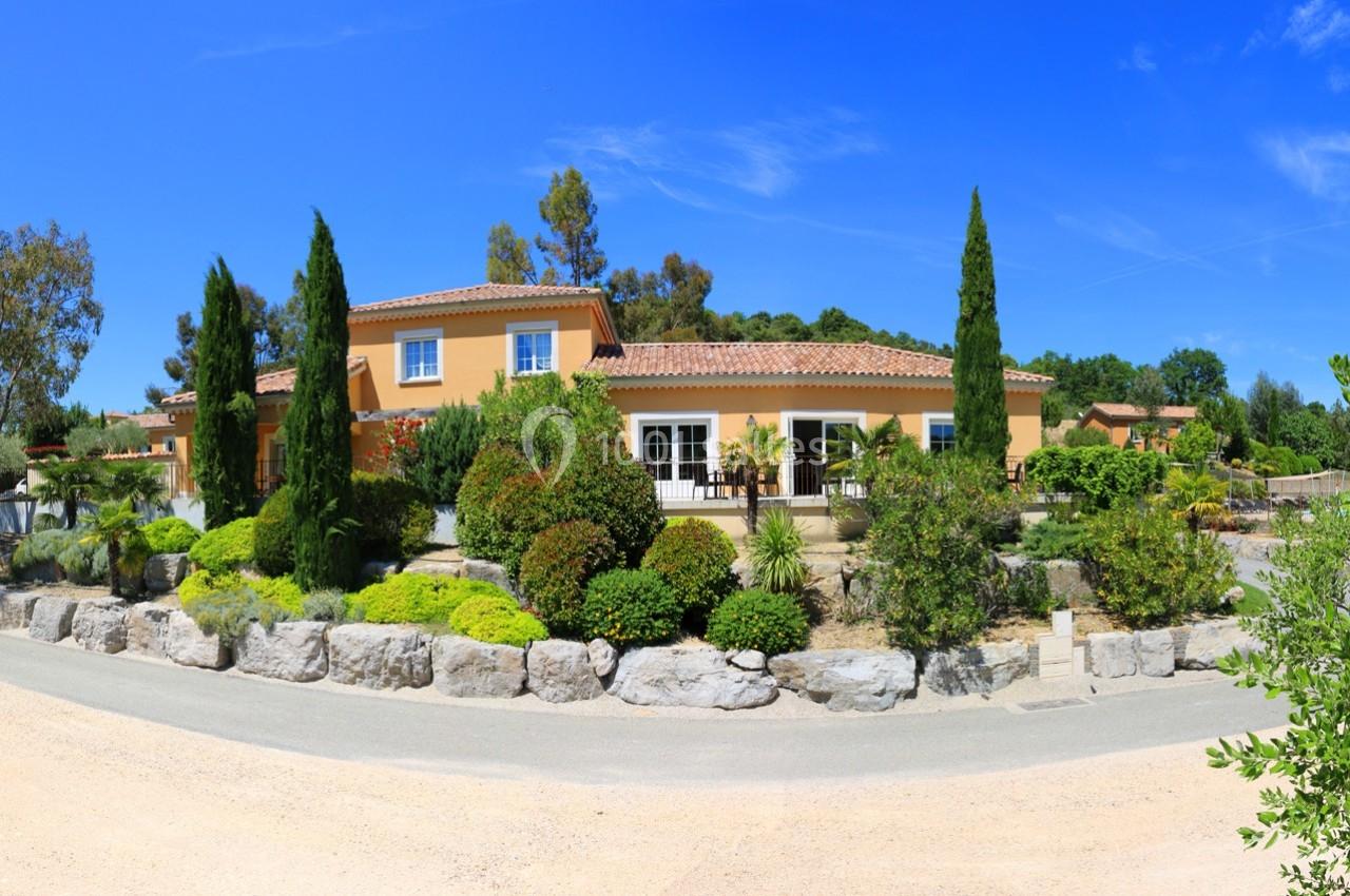 Maison méditerranéenne entourée d'un jardin paysager avec arbres, arbustes et allée en gravier sous un ciel bleu.