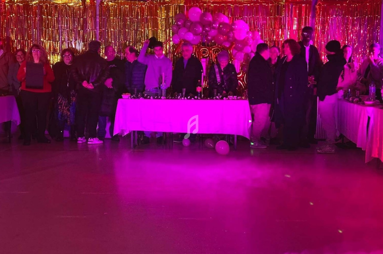 Groupe de personnes debout devant des tables décorées, éclairées par une lumière rose et dorée dans une salle festive.