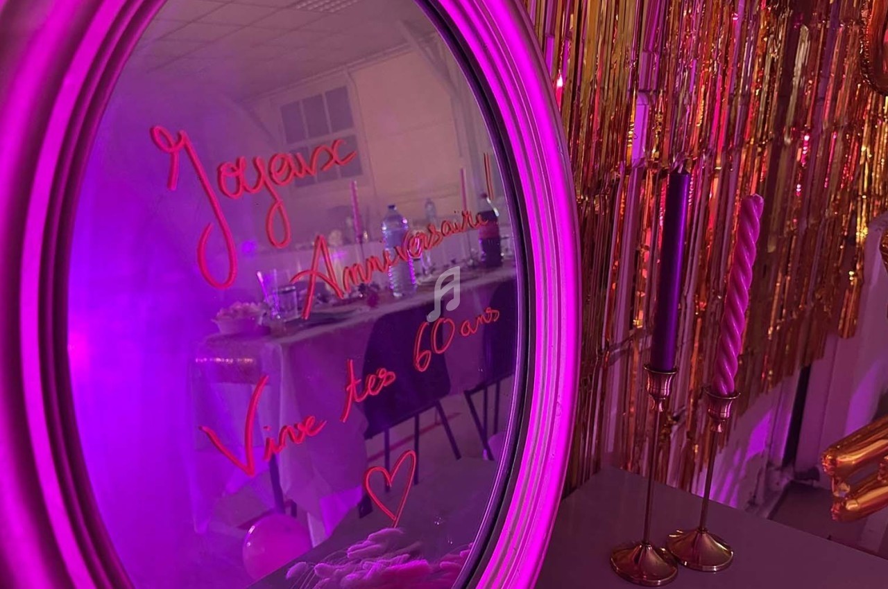 Texte écrit sur un miroir entouré d'une lumière rose, avec des décorations dorées et une table en arrière-plan.