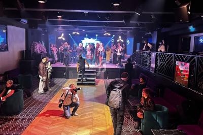 Salle de spectacle avec des personnes sur scène et dans le public, ambiance animée avec éclairages colorés.