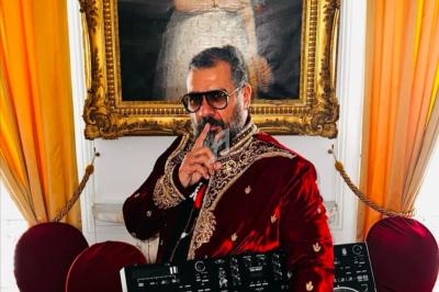 Un homme en tenue élégante tient un micro et une table de mixage dans une pièce ornée de rideaux et d'un tableau.