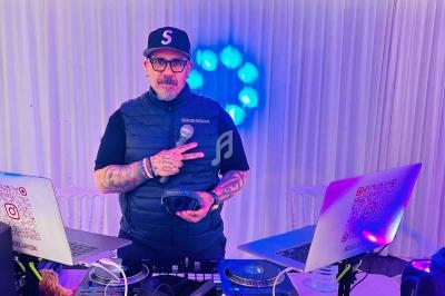 Un DJ debout derrière une table de mixage avec deux ordinateurs portables, tenant un micro et un casque audio.