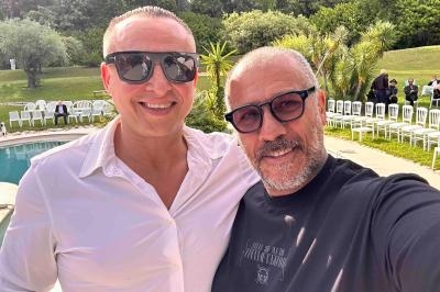 Deux hommes souriants posent pour un selfie près d'une piscine, avec des chaises blanches et un jardin en arrière-plan.
