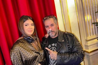 Un homme en veste cloutée et une femme avec un casque autour du cou posent devant un rideau rouge et un sapin décoré.