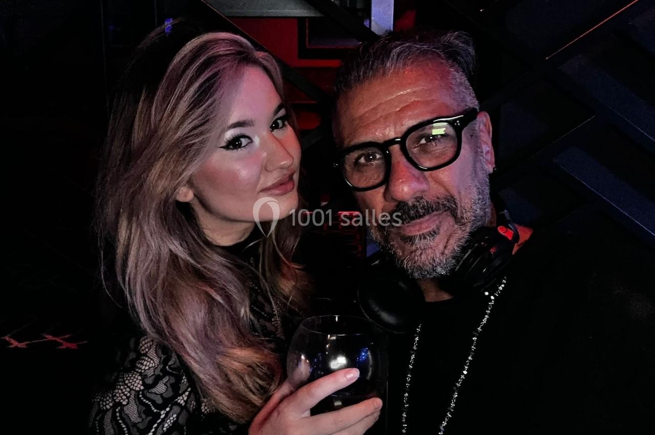Un homme et une femme posent ensemble dans un environnement sombre, la femme tenant un verre à la main.