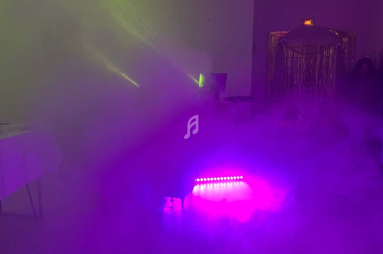 Salle obscure avec éclairage violet et vert, fumée diffuse, table dressée sur la gauche et rideaux brillants en arrière-plan.