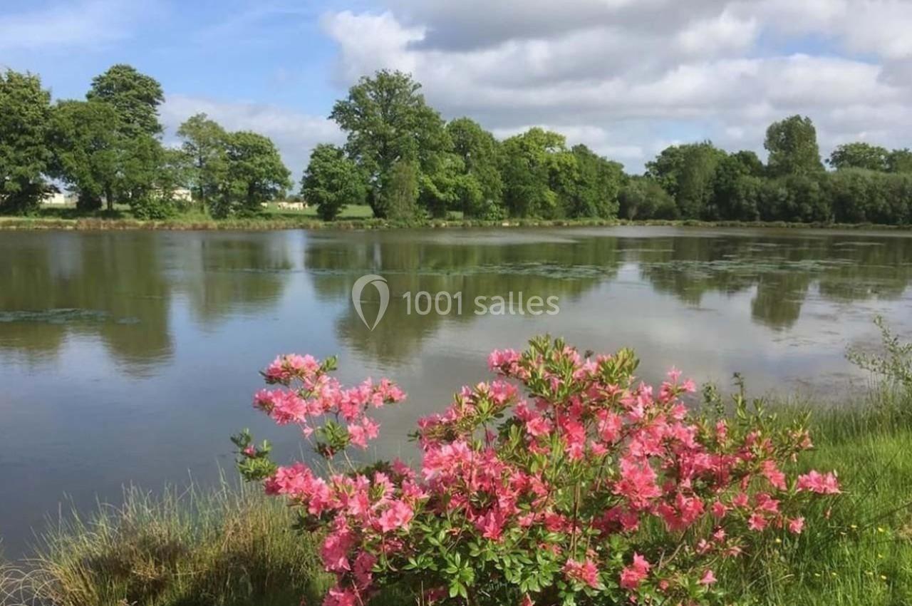 Arbuste fleuri avec des fleurs roses au bord d’un étang entouré d’arbres sous un ciel partiellement nuageux.