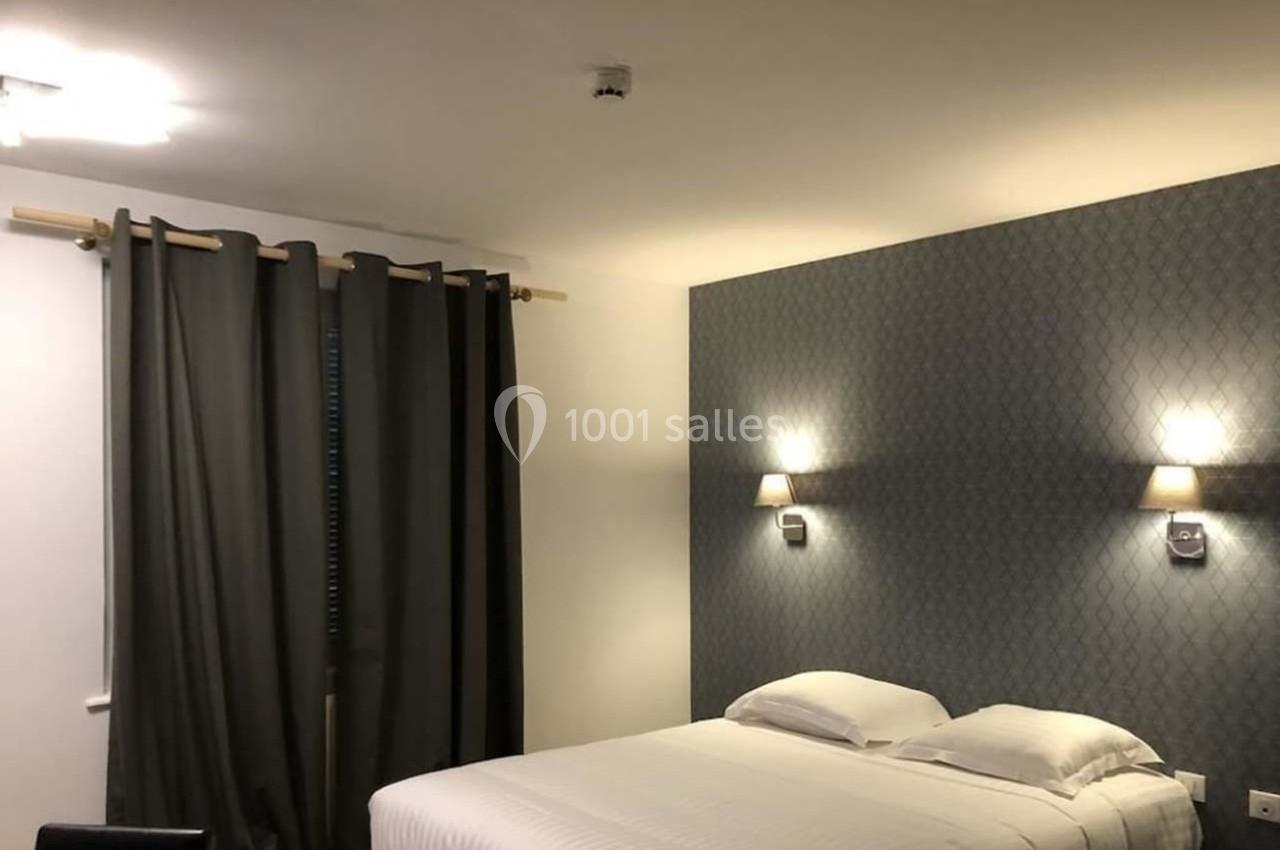 Chambre avec un lit double, des murs décorés de papier peint, des rideaux sombres et deux appliques lumineuses.