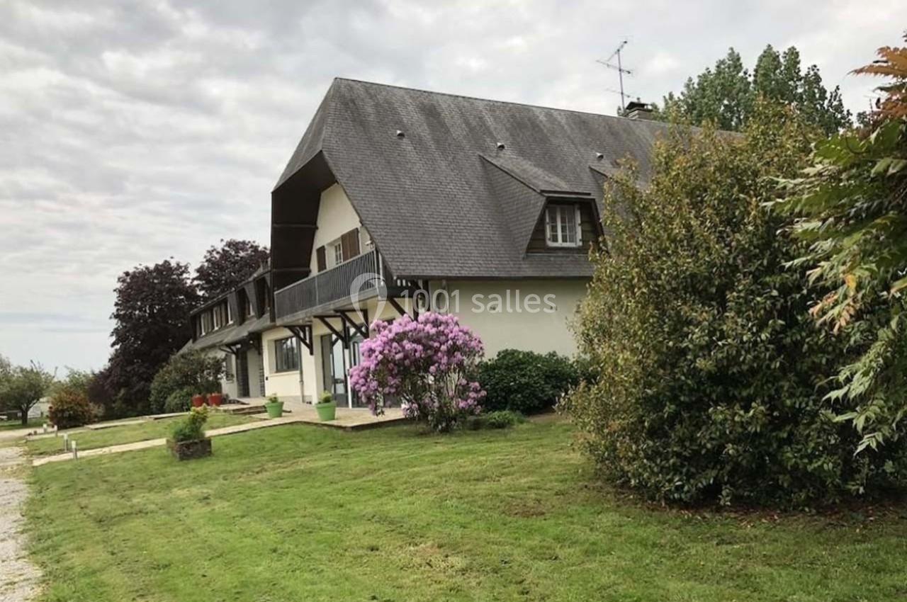 Grande maison avec toit en pente, entourée de pelouse, d'arbustes et d'un arbre fleuri, sous un ciel nuageux.