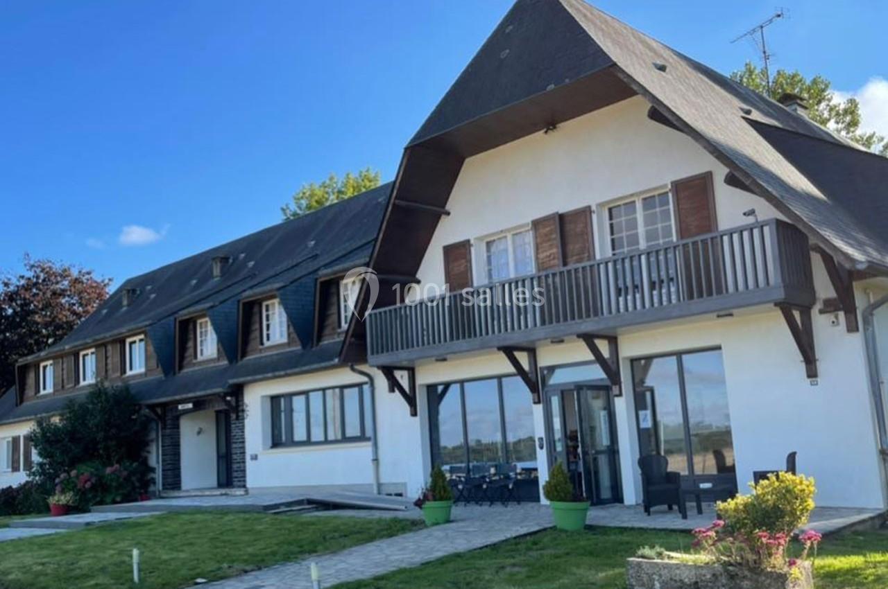 Grande maison à deux étages avec façade blanche, balcons en bois, jardin aménagé et ciel bleu en arrière-plan.