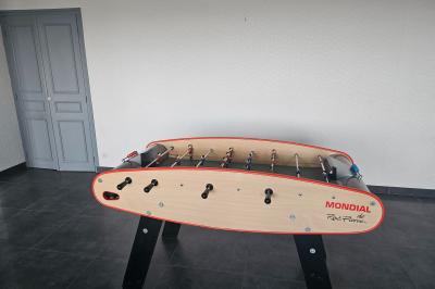 Baby-foot en bois clair avec pieds noirs, situé dans une pièce au sol sombre et murs clairs avec une porte grise.