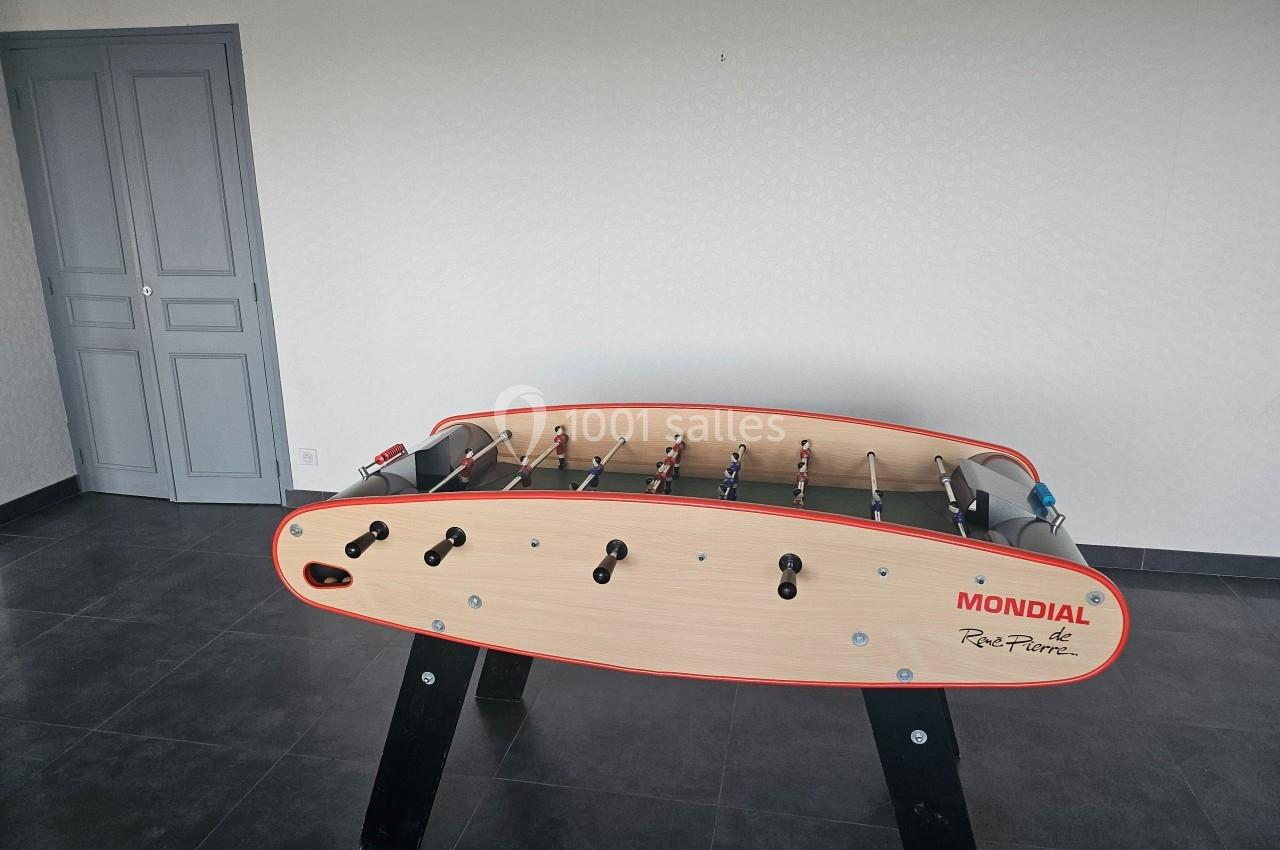 Baby-foot en bois clair avec pieds noirs, situé dans une pièce au sol sombre et murs clairs avec une porte grise.