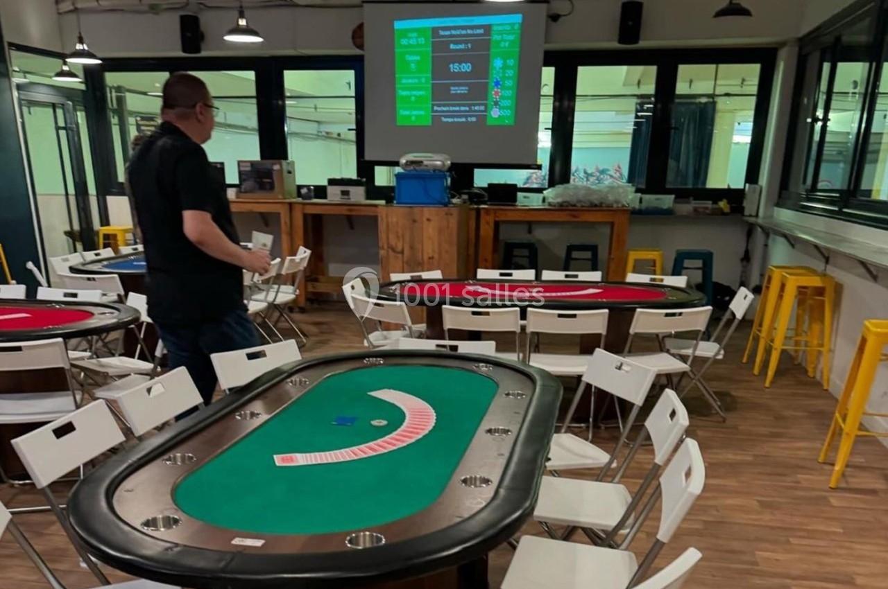 Salle avec des tables de poker, des chaises blanches, un écran de projection et un homme debout près des tables.